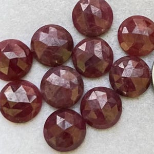 May include: Ten faceted, round, dark red gemstones are displayed. Each gem has a domed, multi-faceted surface. The gems are arranged on a light surface, with a coin for scale. These gems are likely for jewellery making, with a deep red hue.