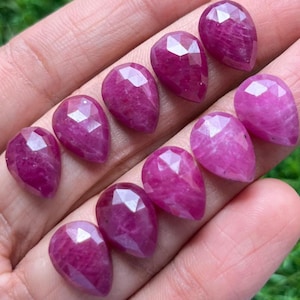May include: A selection of faceted, teardrop-shaped gemstones in shades of deep pink and magenta. The gems are arranged in rows, displaying their polished surfaces and geometric cuts. The gemstones are held in a hand.