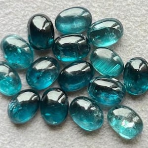 Puede incluir: Una colección de piedras de vidrio lisas, de forma ovalada, de color verde azulado. Las piedras tienen una superficie brillante y reflectante, con un tono verde azulado más oscuro en el centro. Las piedras están dispersas sobre una superficie gris claro.