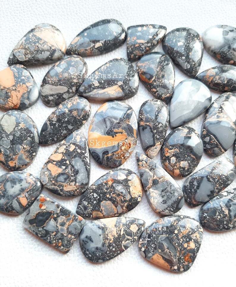 May include: A collection of 25 natural gemstone cabochons in a teardrop shape. The stones are a mix of black, gray, and brown with a speckled pattern. The stones are approximately 1 inch in length.