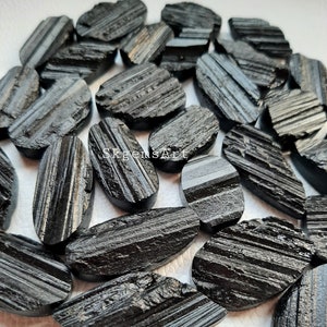 May include: A collection of black, oval-shaped, striated black tourmaline stones. The stones are raw and have a rough, natural texture.