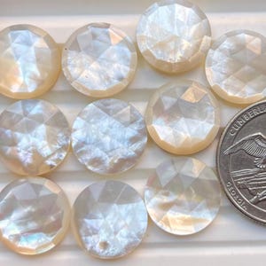 Puede incluir: Una colección de diez cabujones redondos y facetados de nácar iridiscente. Los cabujones son de color blanco crema con un patrón geométrico. Se incluye una moneda como referencia de tamaño.