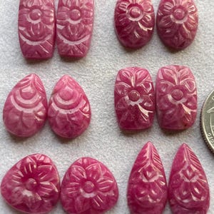 May include: A collection of carved pink gemstones in various shapes, including ovals, rectangles, teardrops, and triangles. Each gemstone features a floral design. The gemstones are displayed on a light gray surface.