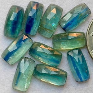 Puede incluir: Un conjunto de nueve piedras preciosas de cianita rectangulares y facetadas que muestran un degradado de tonos azul verdoso translúcido, verde claro y azul intenso. Cada piedra presenta una superficie pulida con facetas geométricas.