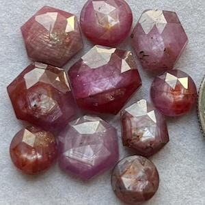 May include: A selection of faceted, reddish-pink gemstones. The gems are in various shapes, including round and hexagonal forms. Each stone has multiple facets that reflect light, creating a sparkling effect. The gemstones are arranged closely together, showcasing their colour variations.