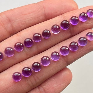 Könnte beinhalten: Eine Sammlung von 18 runden, violetten Amethyst-Cabochons, jeder etwa 6 mm im Durchmesser. Die Cabochons sind in zwei Reihen auf einer Hand angeordnet.