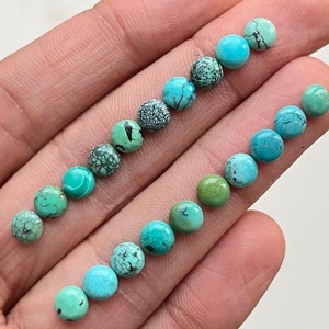 May include: A collection of 15 round turquoise cabochons in various shades of blue and green. The cabochons are arranged in a line on a hand.