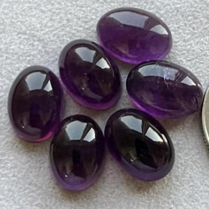 May include: Six polished, oval-shaped amethyst gemstones. The gemstones are a deep purple color with a glossy, reflective surface. The stones are arranged on a light gray surface.