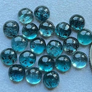 May include: A collection of 20 round, blue-green gemstones with black flecks. The gemstones are smooth and polished, and they are arranged in a random pattern.