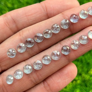 May include: A collection of 18 faceted aquamarine gemstones, each with a round shape and a light blue colour. The gemstones are arranged in two rows on a hand.