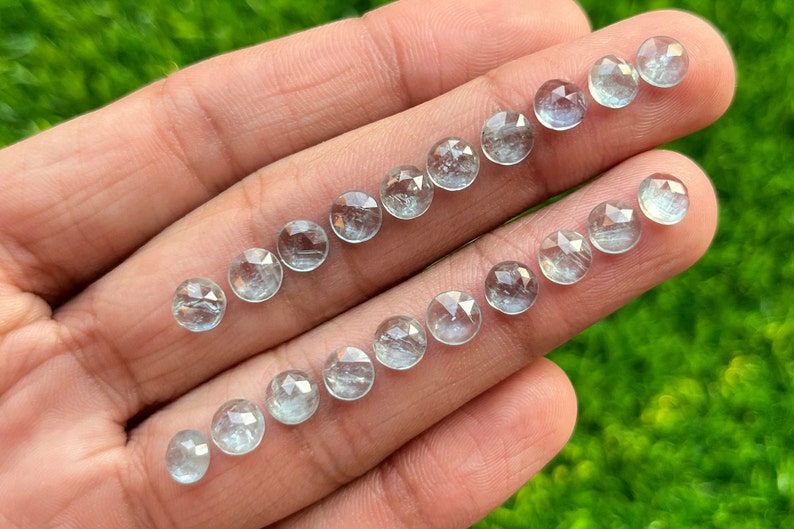 May include: A collection of 18 faceted aquamarine gemstones, each with a round shape and a light blue colour. The gemstones are arranged in two rows on a hand.