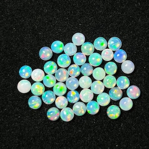 May include: A collection of round, iridescent opal gemstones in various shades of white, blue, and green. The gemstones are arranged in a loose pile on a black background.