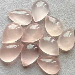 Könnte beinhalten: Eine Sammlung von neun polierten Rosenquarz-Edelsteinen in Tropfenform. Die Steine variieren in Rosatönen und sind durchscheinend mit glänzender Oberfläche. Sie liegen auf einer hellgrauen Oberfläche, die die natürliche Schönheit der Edelsteine hervorhebt.