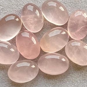 Könnte beinhalten: Eine Sammlung von neun ovalen Rosenquarz-Edelsteinen. Die polierten Steine variieren in Rosatönen und haben eine glänzende, reflektierende Oberfläche. Die Edelsteine liegen auf einer hellgrauen Oberfläche.