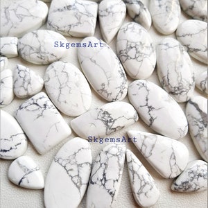 May include: A collection of white howlite cabochons with gray veining. The stones are various shapes and sizes, including oval, teardrop, and rectangular. SkgemsArt is printed on the image.