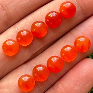 Puede incluir: Diez cabujones redondos de cornalina naranja, cada uno con una superficie lisa y pulida. Los cabujones están dispuestos en dos filas de cinco, con la fila superior ligeramente desplazada de la fila inferior.