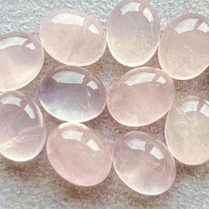 Könnte beinhalten: Eine Sammlung von zehn ovalen Rosenquarz-Cabochons. Die polierten Steine variieren in Rosatönen und liegen auf einer hellen Oberfläche. Die glatten, durchscheinenden Edelsteine sind eng beieinander angeordnet.