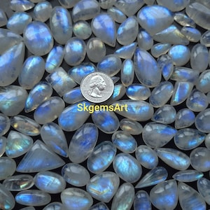 May include: A collection of iridescent moonstone cabochons in various sizes and shapes. The stones are a milky white with a blue sheen. A US quarter is included for scale.