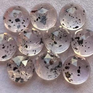 Könnte beinhalten: Eine Sammlung klarer, facettierter Edelsteine mit schwarzen Einschlüssen. Die runden Steine haben ein poliertes Aussehen und sind eng beieinander angeordnet. Die Edelsteine sind wahrscheinlich für die Schmuckherstellung oder das Basteln gedacht.