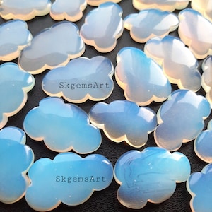 May include: A collection of iridescent opal cloud-shaped cabochons. The cabochons are a light blue colour with a white iridescent sheen. The cabochons are arranged in a random pattern on a black surface. The text "SkgemsArt" is visible on two of the cabochons.