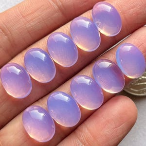 May include: Ten oval-shaped, iridescent gemstones with a light purple and pink hue. The gemstones are arranged in a row on a hand.