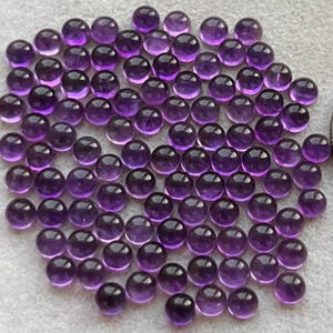 May include: A collection of round, polished amethyst gemstones. The gemstones are a deep purple color, with a smooth, reflective surface. The gemstones are scattered across a light gray surface.