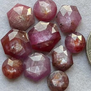 Könnte beinhalten: Eine Sammlung facettierter, rötlich-rosa Edelsteine in verschiedenen Formen, darunter rund und sechseckig. Die Edelsteine haben eine polierte, reflektierende Oberfläche, die innere Muster und Farbvariationen zeigt. Die Anordnung befindet sich auf einer hellen Oberfläche.