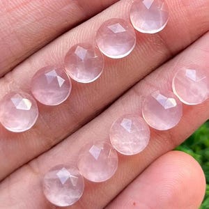 Könnte beinhalten: Zehn facettierte Rosenquarz-Cabochons in verschiedenen Größen, von hellrosa bis dunkelrosa, auf einer Hand präsentiert.