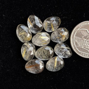 May include: Nine clear quartz stones with gold and brown inclusions, faceted and oval shaped. The stones are arranged in a circle on a black surface. A UK 20 pence coin is included for size reference.