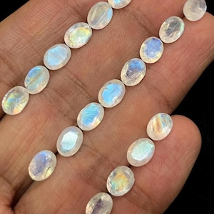 Puede incluir: Gemas de piedra lunar ovaladas con un brillo nacarado y destellos internos azules y amarillos. Las gemas están dispuestas en filas, mostrando sus superficies pulidas. Las piedras se sostienen en una mano.