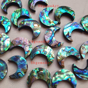 May include: A collection of 18 iridescent abalone shell crescent moon shaped cabochons. The cabochons are approximately 2.5 cm in length and have a variety of colours, including green, blue, purple, and gold.