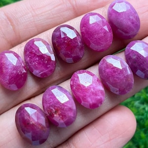 May include: A collection of oval-shaped, faceted rubies in varying shades of pink and red. The gemstones are arranged closely together, showcasing their polished surfaces and internal clarity. The rubies are displayed against a neutral background.