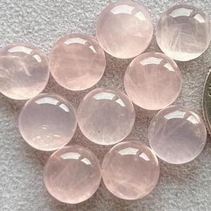 Könnte beinhalten: Zehn polierte Rosenquarz-Cabochons in sanftem Rosa und transluzenter Optik. Die runden Edelsteine liegen auf einer hellgrauen Oberfläche. Eine Münze dient als Größenvergleich. Jeder Cabochon hat einen Durchmesser von etwa 2,5 cm.