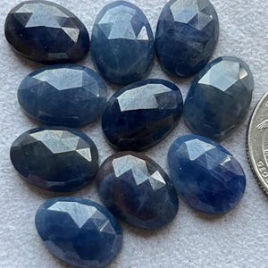 Puede incluir: Colección de nueve zafiros facetados de forma ovalada en varios tonos de azul. Las piedras preciosas tienen una superficie pulida y reflectante, con facetas visibles que captan la luz. Los zafiros están dispuestos sobre una superficie gris claro.