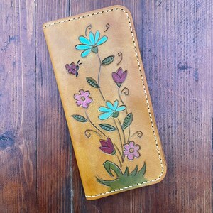 Op de afbeelding: Een bruine leren portemonnee met een bloemenontwerp. De bloemen zijn blauw, roze en groen. De portemonnee heeft een gestikte rand.