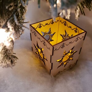 May include: A wooden candle holder with a square shape and intricate cut-out designs of suns. The candle holder is lit and glows a warm yellow light.