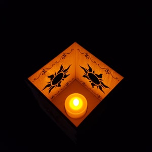 May include: A wooden box with a candle inside. The box is decorated with a black silhouette of the sun with rays. The box is lit from inside by the candle.