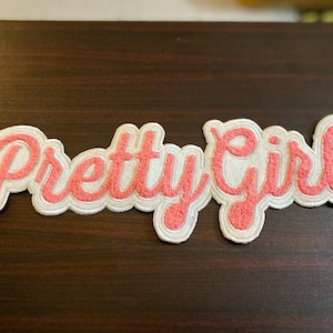May include: A pink and white felt patch with the text "Pretty Girl" in a cursive font.