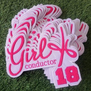 May include: A stack of pink felt patches with the word "Girl" in pink cursive lettering and the word "conductor" below it. The number "18" is in pink felt below the words.