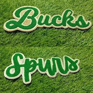 Puede incluir: Dos parches bordados con texto verde sobre un fondo dorado y blanco. El parche superior dice "Bucks" en una fuente de escritura, y el parche inferior dice "Spurs" en un estilo similar. Los parches están colocados sobre un fondo de césped artificial verde.