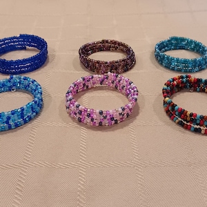 May include: Six multi-row wrap bracelets made of small glass seed beads. The bracelets feature various color combinations including solid royal blue, dark brown, turquoise, a blue gradient mix, a pink and purple mix, and a multi-colored earth tone pattern.