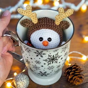 a person is holding a cup with a crochet snowman with antlers in it
