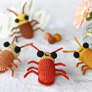 May include: Handmade crochet insect toys in various shades of orange, brown, and yellow. Each insect has black eyes and antennae. The toys are arranged on a white surface, with a few flowers in the background.