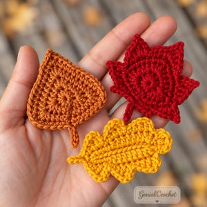 May include: Three crocheted leaf appliques in autumn colours: one orange triangular leaf, one red maple-shaped leaf, and one yellow oak-shaped leaf. The text Genial Crochet is visible in the bottom right corner of the image.