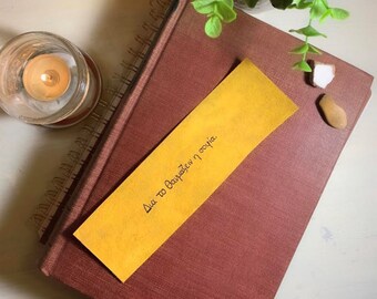May include: A yellow bookmark with the text "Για το δανεισμένο η σοφία." written in black ink. The bookmark is placed on top of a brown leather-bound book.
