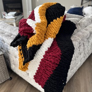 May include: A chunky knit throw blanket with stripes of burgundy, white, black, and mustard yellow.