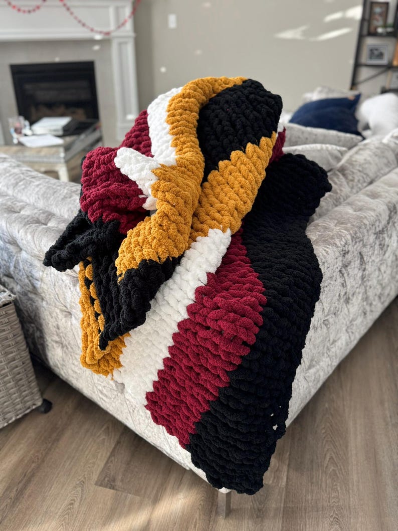 May include: A chunky knit throw blanket with stripes of burgundy, white, black, and mustard yellow.