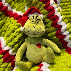 May include: A plush green character wearing a red Santa hat with white trim, sitting on a lime green, red, and white knitted blanket. The character has yellow eyes and a red heart on its chest. The blanket has a circular pattern.