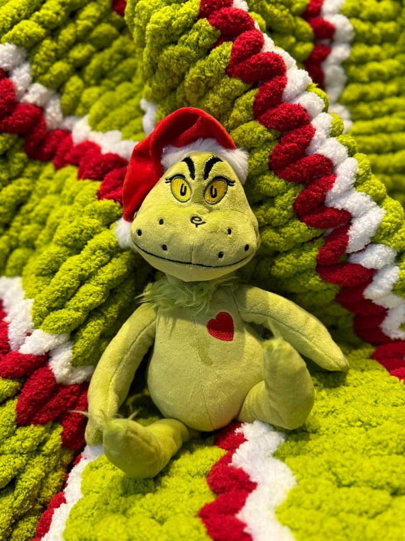 May include: A plush green character wearing a red Santa hat with white trim, sitting on a lime green, red, and white knitted blanket. The character has yellow eyes and a red heart on its chest. The blanket has a circular pattern.
