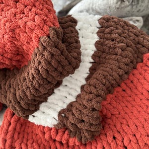 K&ouml;nnte beinhalten: Eine grob gestrickte Decke in Rost-, Braun- und Wei&szlig;t&ouml;nen. Die Decke hat dickes, strukturiertes Garn, das ein gem&uuml;tliches und einladendes Aussehen erzeugt. Das Farbschema umfasst eine rostfarbene Basis mit braunen und wei&szlig;en Streifen.